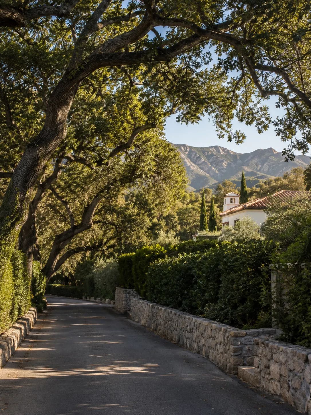 An oak-lined lane in Montecito with a stone wall, red-tile roof, and mountains beyond.